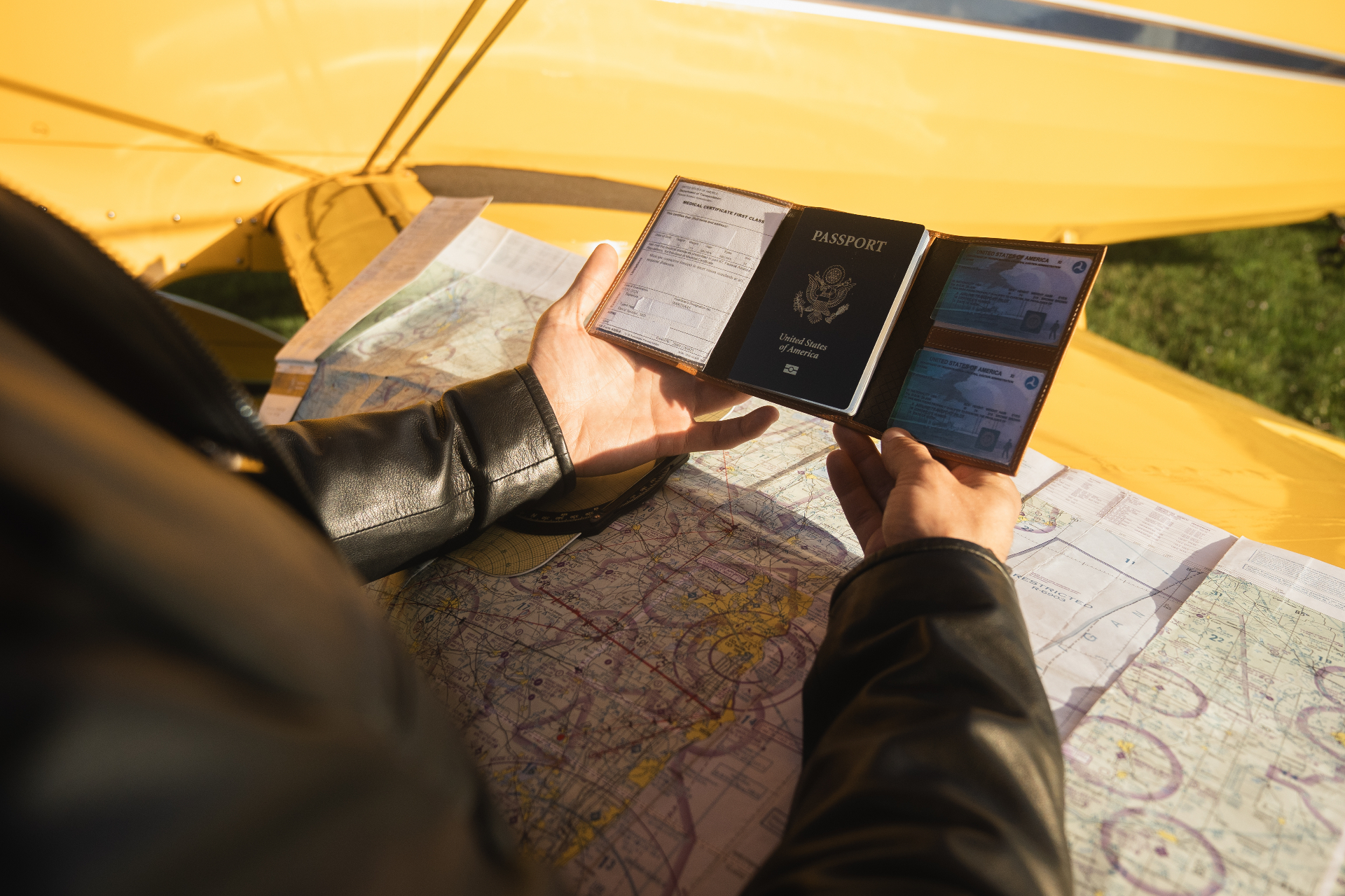 Pilot in the cockpit holding an open leather passport wallet with US passport and pilot licenses over an aviation sectional chart, yellow aircraft framing the scene